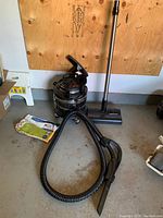 Full view of the vacuum unit with hose, metal wand, floor attachment, and new filter packet on the floor, showing all main components.