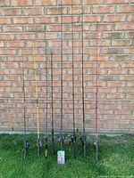 Eight used fishing rods with reels attached standing upright against a brick wall outdoors on grass.