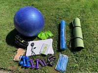 Full lot with yoga ball, weights, mats, helmet, bands, and stopwatch laid out outdoors on grass