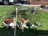 Front wide shot of all gardening items including plant stands, pots, tools, lantern and accessories arranged on grass in sunlight.