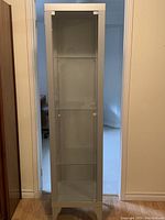 Front view of tall narrow glass cabinet with metal frame and three glass shelves inside.