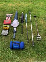 All golf accessories laid out on grass including umbrellas, golf balls, ball retriever, hole maker, and blanket