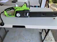 Side view of Greenworks electric chainsaw body and protective bar cover on white folding table.