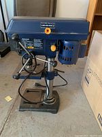 Front-left angled view of the Mastercraft 8-inch drill press showing the motor housing, three-spoked feed handles, and base with power cord.