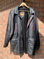 Black Old Hide House leather coat displayed on hanger against brick wall showing front, collar, pockets, and maroon accents.