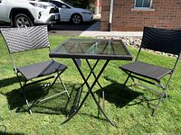Full view of outdoor patio bistro set on grass showing glass top square table and two folding chairs with woven rattan design.