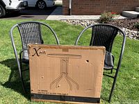 Photo of two black wicker chairs with curved metal frames standing on grass with the boxed glass top table in front showing the brand 'Xeilla'.