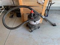 Shop-Vac 10-gallon wet/dry vacuum with hose and floor attachment on concrete floor next to wooden shelves.