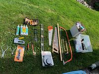 Top-down view of various used hand tools and hardware including handsaws, caulking tubes, staple gun, screwdrivers, measuring tape, light bulbs, storage bins, and flashlight spread on grass.