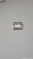 Close-up photo of single emerald cut rose quartz gemstone showing rectangular shape with step cuts and light reflection on polished facets, placed on plain white background.