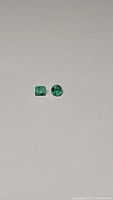 Photo of two emerald gemstones, one emerald cut and one round cut, placed side by side on a plain surface.