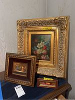 Photo showing three different framed still life paintings set on a surface with visible size tags on the frames (19x20, 7x3, 3x7). Frames are decorative gold-toned with different framing styles. Paintings depict flowers and fruit.