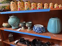 Shelf with nesting dolls, lidded urn, cups, turquoise planter, and blue speckled planter base.
