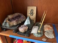 Shelf display with two amethyst geodes, stone pyramid, Chinese character slab, pen set, and loose stones.