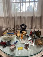 Overview photo showing multiple animal figurines including owl, ram, lion, dog, turtle, birds; metallic bells, a large metal apple, and metal gear plaques on a glass table by a window