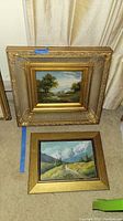 Two framed landscape paintings positioned on floor against curtains, showing ornate gold frame for the countryside scene and simpler gold frame for mountain valley.