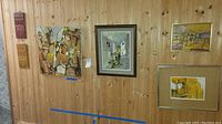 Six original artworks hung on wood paneled wall, combination of abstract and architectural scenes, various frame types.