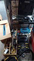 Photo showing workshop tools including wooden tool box with hardware, hand tools, Dewalt drill charger below, and various bits and screwdrivers laid out on shelf.