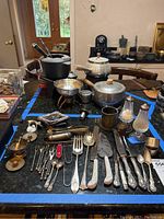 Overview photo showing all silverware items including utensils, cups, salt and pepper shakers, tongs, napkin rings, and cake cutter on black surface with blue tape border.