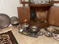 Multiple silverplate trays, a silverplate pedestal bowl, and charger plates along with wooden, ceramic and glass platters arranged on a tiled floor.