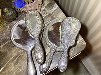 Three ornate handheld mirrors with beveled glass and detailed silver-plated handles and backs