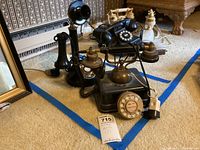 Two vintage telephones displayed on carpeted floor near furniture showing the candlestick style rotary dial phone and classic black rotary dial desk phone.