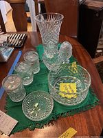 Overview of eight crystal pieces arranged on a green cloth on a wooden table: tall vase, covered jar, two bowls, four smaller containers, and a small dish.