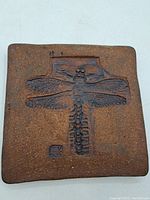 Front view of square ceramic tile with embossed dragonfly design and artist mark.