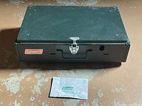 Closed green metal Coleman folding camping stove with latch and Coleman sticker on front, laying on floor.