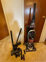 Dirt Devil upright vacuum with many attachments laid out on a carpeted floor against a wooden door background.