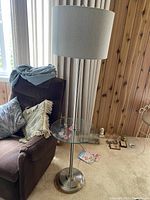 Front and side view of floor lamp showing brown cushioned chair nearby and round glass table integrated in lamp stand.