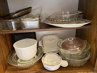 Photo showing various clear glass casserole dishes stacked on wood shelf including rectangular and round shapes, also white ceramic mixing bowl and textured glass bowls.
