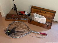 Photo showing fishing rod with reel standing against wall, wooden tacklebox with compartments filled with fishing lures, bobbers, and hooks, fishing net with metal frame and red handle, and some accessories like fishing line and boxes.