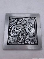 Front view of metal plaque with stylized raven crest design in black and silver tones.