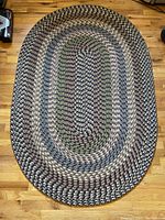 Top-down photo showing the entire oval braided rug with concentric rings of various earth tones on a wooden floor.