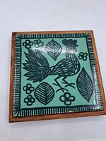Front view of ceramic folk art tile featuring black bird and leaf design on green background in wooden frame.