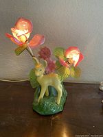 Front view showing the vintage ceramic yellow deer lamp with two lit bulbs covered by pink flower-shaped shades, surrounded by green leaves and pink flowers on a green base.