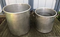 Two large aluminum commercial cooking pots shown side by side on an outdoor surface, the bigger one significantly taller and wider than the smaller one.