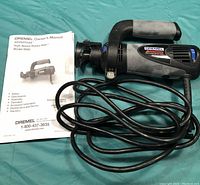 Top view of the Dremel 9000 rotary saw with power cord arranged, owner's manual card placed beside it on a green surface.