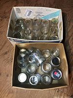 Two boxes filled with assorted clear glass mason jars, various sizes and brands, some with lids and some without