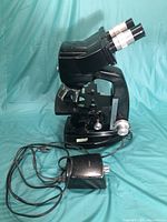Side view showing the black body of the microscope, silver eyepieces, objective lenses, and electrical cord attached to the external light source.