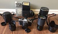 Photo showing Pentax K-1000 camera body with attached lens, two additional lenses with lens hoods, lens cap, and Sunpak 383 flash unit with box all arranged on wood floor