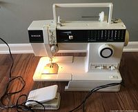 Full view of the white Singer sewing machine Model 7028 showing the sewing area, foot pedal, power cord, and built-in light illuminated.