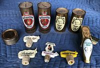 Four vintage Ballantine beer cans (two coin bank style with slots on top, two display style), pile of metal lids, Ballantine tap handle box, and four liquor pourers with corks arranged on blue surface.
