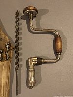Vintage metal hand brace with wooden handle and rotating head cap, lying on flat surface with one large auger bit beside it.