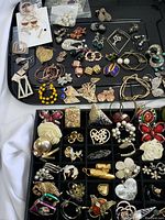 Black tray with various pairs of earrings, brooches, and pendants arranged displaying diverse styles, some with pearl and rhinestone accents.