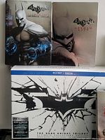 Photo showing two Batman Arkham City steelbooks with Batman and glowing fists artwork and the Armored Batman steelbook in a metal casing, along with the Dark Knight Trilogy Ultimate Edition Blu-ray set with shattered bat symbol on white box.