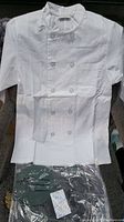 Full view of a white Chef Works chef jacket with double-breasted button design and front pocket, laid out on a chair.