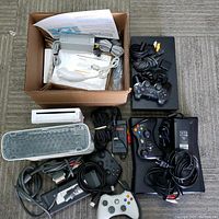 Top-down view showing two black Xbox 360 consoles, one white Xbox 360 controller, assorted cables, one grill-pattern sensor bar accessory and a cardboard box of Wii accessories.