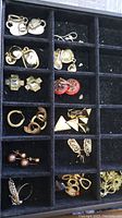 Close-up photo showing various pairs of gold-tone and silver-tone earrings in a compartment organizer box including hoop earrings, clip-ons, and decorative styles
